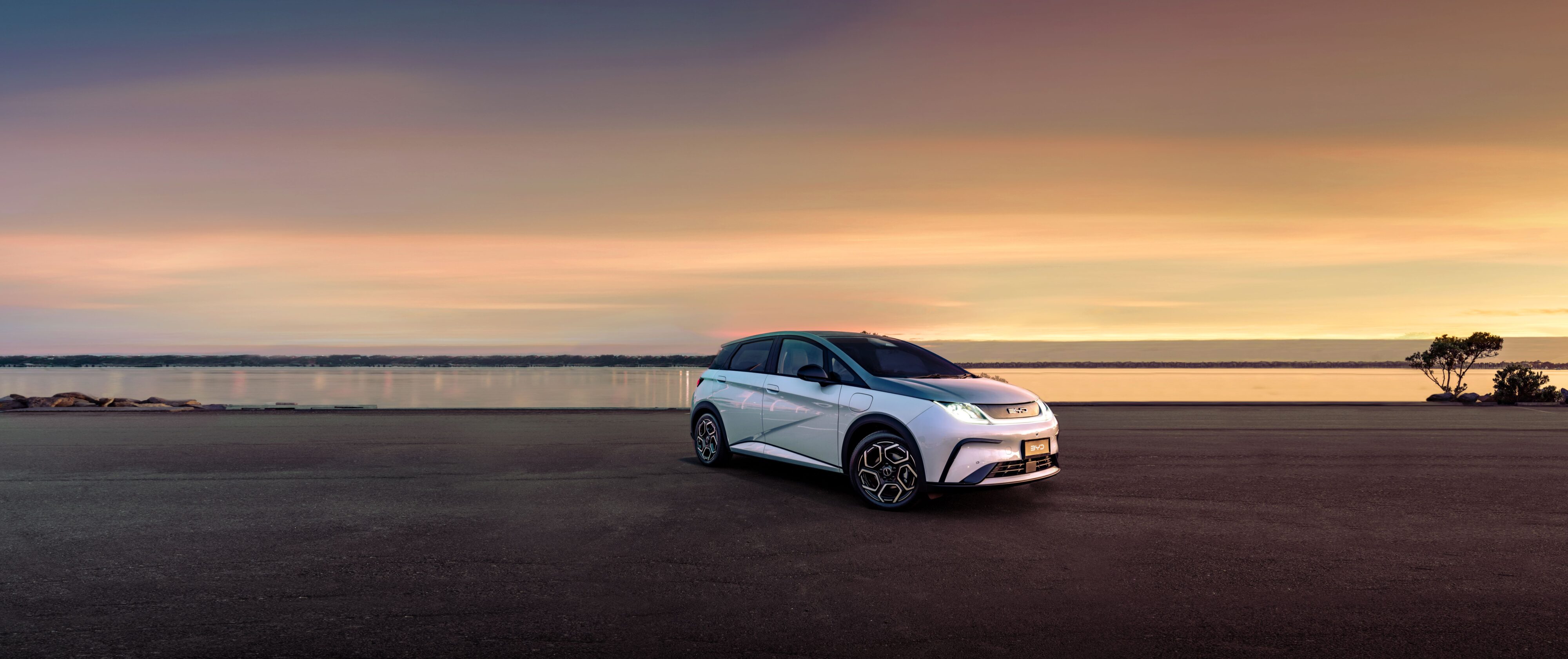 Front three quarters view of a BYD Dolphin in white with a contrast black roof. Backdrop is of a coast and photo was taken at sunset.