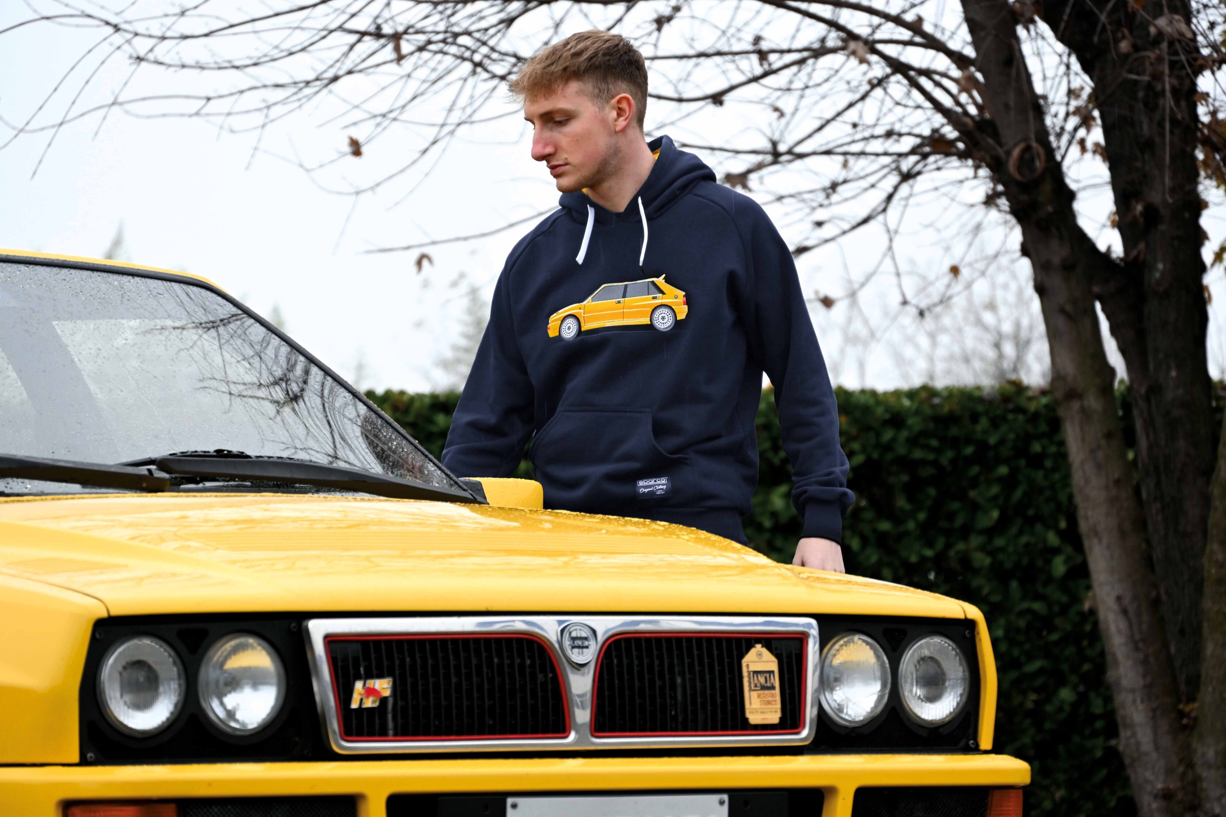 A model in a Sparco Lancia Delta Integrale sweater poses next to a Lancia Delta Integrale HF in yellow.