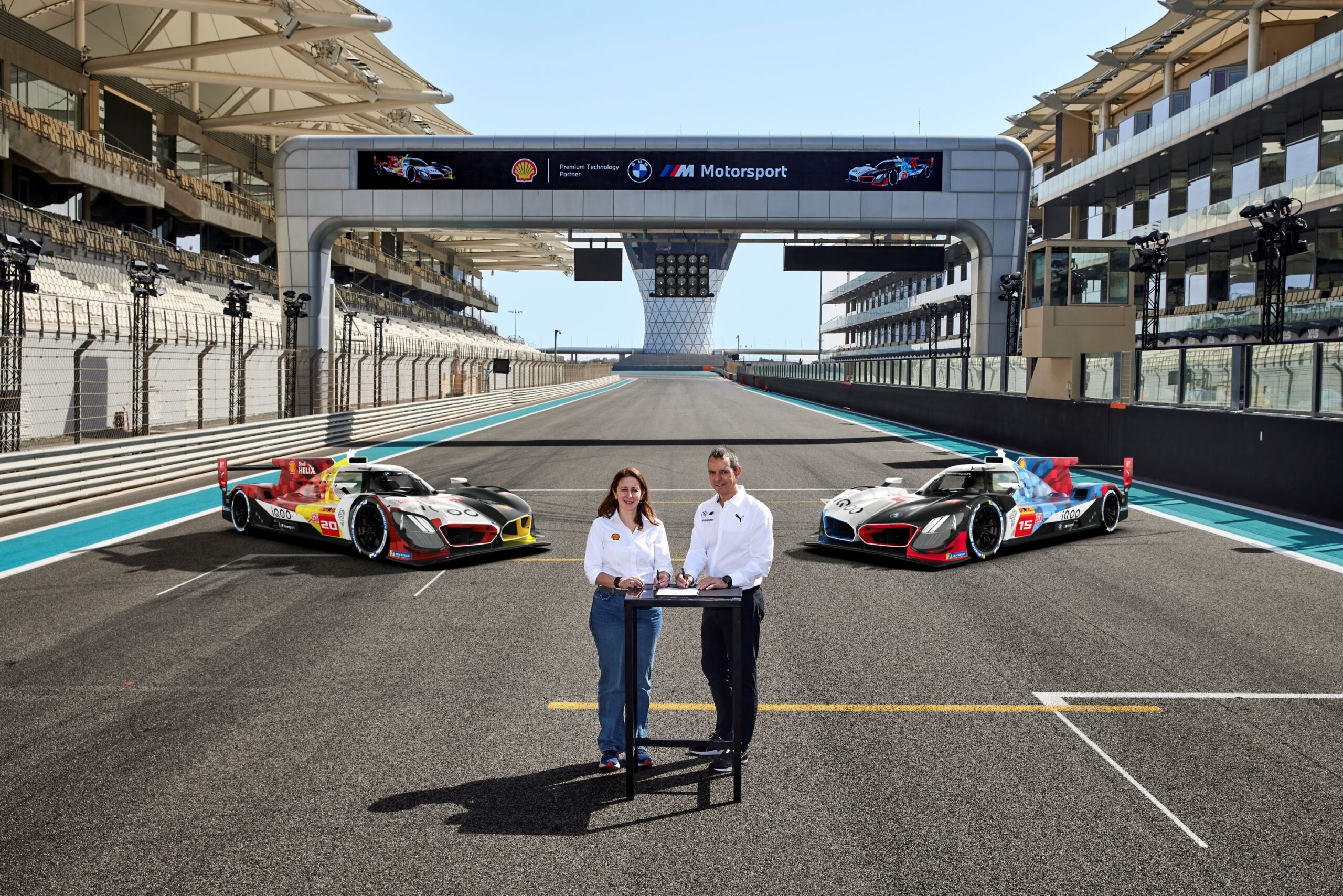 Aysun Akik of Shell Lubricants and Franciscus van Mell of BMW M Motorsport signing a new partnership agreement on a racetrack with two BMW Shell WEC cars in the background.