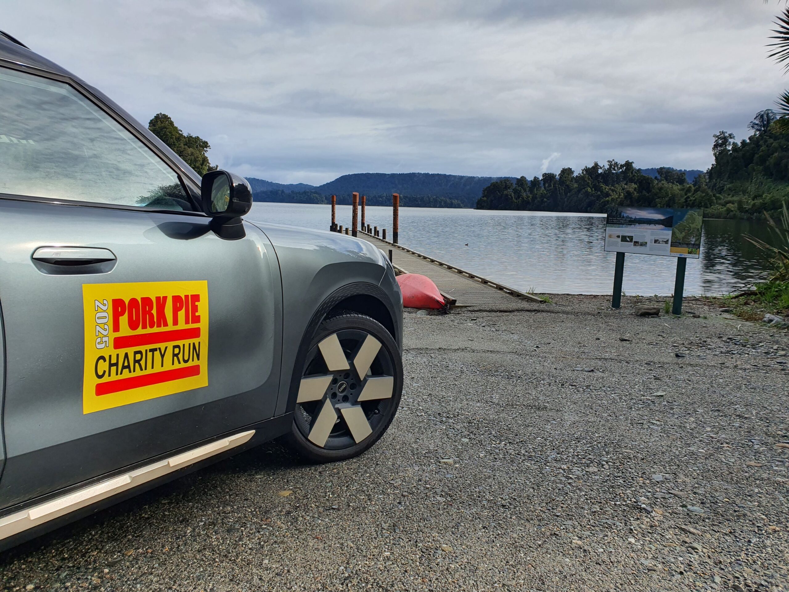 A photo of the right side of a 2025 Mini Countryman SE All4 with a 2025 Pork Pie Charity Run sticker on it. Pictured in the background is Lake Ianthe in the Ross region of New Zealand. Taken on the 2025 Pork Pie Charity Run.