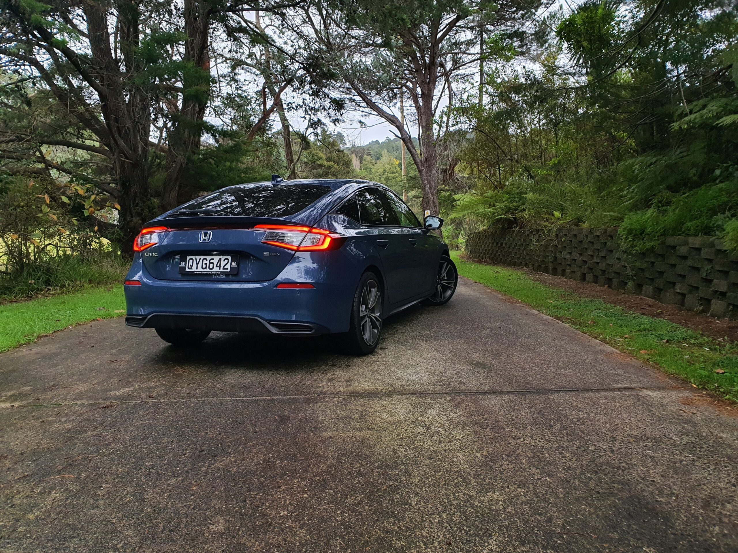 Rear three quarters view of a 2025 Honda Civic e:hev in Ocean Blue Pearl with greenery in the background.
