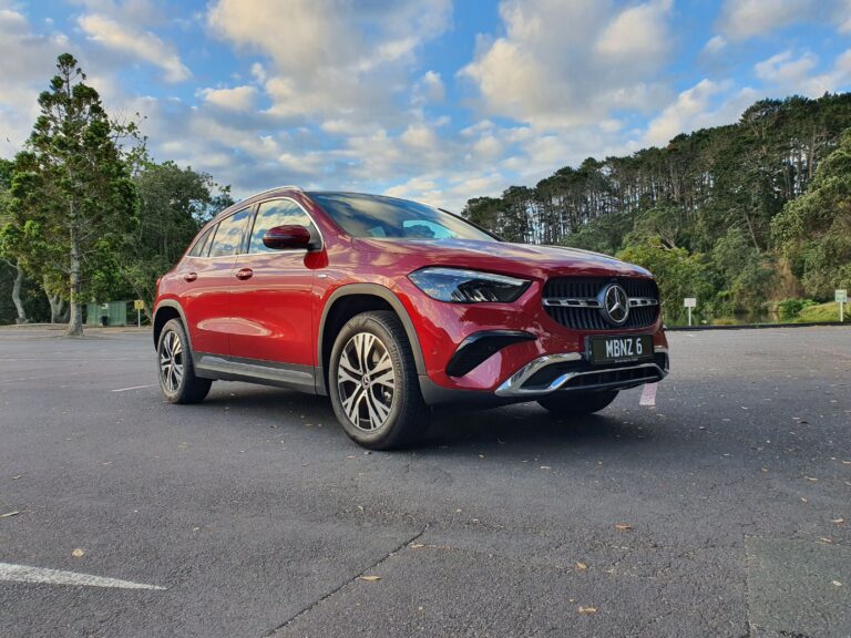 Front three quarters view of a 2025 Mercedes-Benz GLA250e in Patagonia Red Metallic with trees in the background.